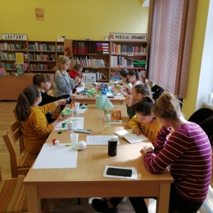 Dzieci z Ukrainy podczas spotkania w bibliotece. Część dzieci przygotowuje prace plastyczne, a dziewczynka z pomocą wolontariuszki uczy się czytać po polsku